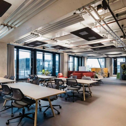 Spacious office to rent at Rosenheimer Straße 143C, featuring modern furnishings and several desks under industrial ceilings, perfect for collaborative work. Bright windows enhance the inviting workspace.