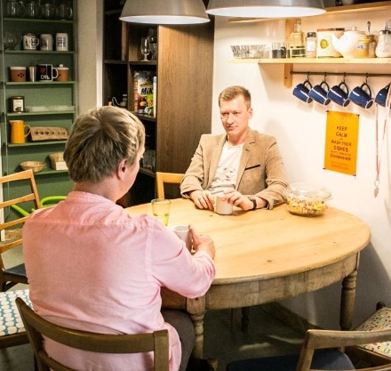 Two people are seated at a wooden table in a cozy, furnished office space, engaging in a conversation. The office includes shelves with cups and decorative items, available for office space rental.