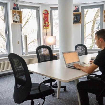 Man working on a laptop in a Munich Giesing office space, featuring a round table, several chairs, and decorated with skateboards by the windows.