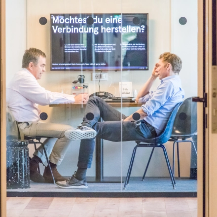 Two people engaged in a discussion in a furnished office space with a digital screen on the wall in Oskar-von-Miller-Ring 20, Munich Maxvorstadt. Ideal for office space rental.