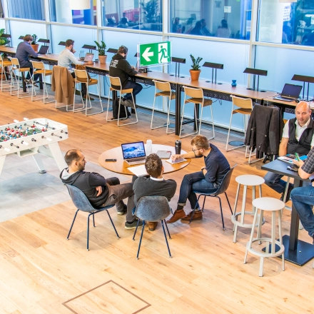 Bright, furnished workspace with people collaborating at tables, featuring long desks and a foosball table. Ideal for rent office space in Munich's Maxvorstadt neighborhood.