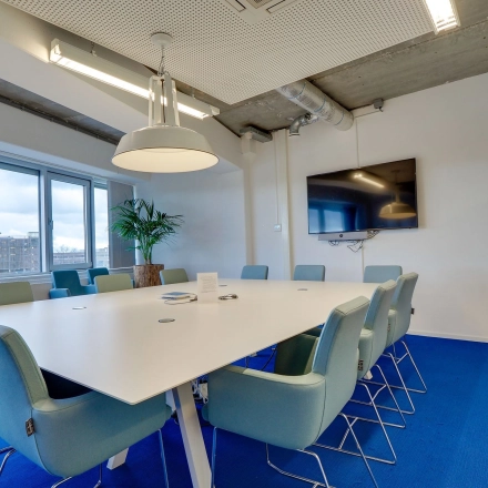 Modern conference room with a large white table surrounded by blue chairs, wall-mounted TV, and a window view at Zoomstede 19, Nieuwegein. Ideal for office space rental.