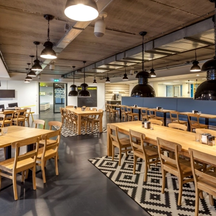 Spacious and modern interior with multiple wooden tables and chairs, perfect for collaborative work or dining. Bright overhead lighting and industrial-style decor add character to this inviting workspace. Ideal for office space rental.