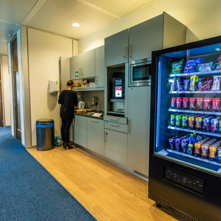 A furnished office space rental in Nijmegen featuring a pantry area with a person preparing a drink beside a stocked vending machine.
