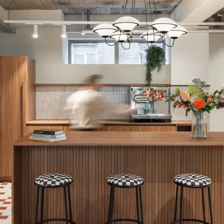 Stylish office lounge area with modern wooden furniture, art on the walls, and a person making coffee at the counter.