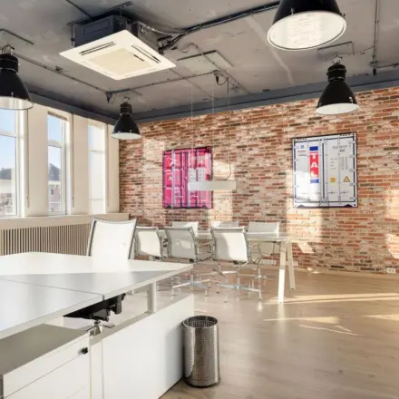 Spacious office space at Veerhaven 7, Rotterdam Center, featuring exposed brick walls, ample natural light, and modern furnishings. Ideal for office space rental with a mix of desks and a meeting area.