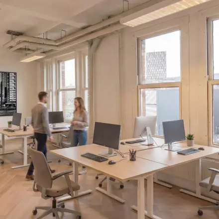 Bright office space with modern desks and computers, situated in Rotterdam Delfshaven. Two people are discussing at a desk. Ideal for office space rental.