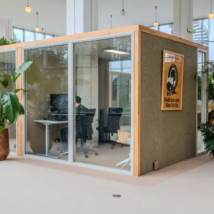 Office space rental at Van Nelleweg 1, Rotterdam Delfshaven. A person works at a desk inside a glass-walled office unit, surrounded by plants and modern furnishings.