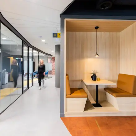 Modern office interior at Wilhelminaplein 1-40, Rotterdam Kop van Zuid, featuring a cozy booth seating area and individuals engaged in business activities in the background. Ideal for office space rental.