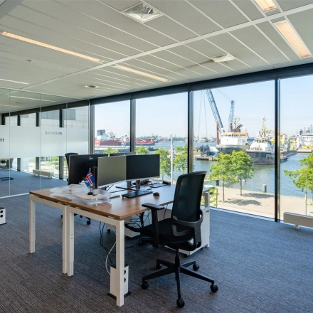 Bright office space with large windows overlooking Rotterdam Waalhaven, featuring desks and chairs for rental. Ideal workspace with modern amenities for business professionals.