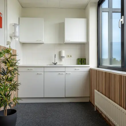 Compact and bright office pantry with wood paneling, modern white cabinetry, and a large window, ideal for office space rental at Curieweg 9, Spijkenisse.