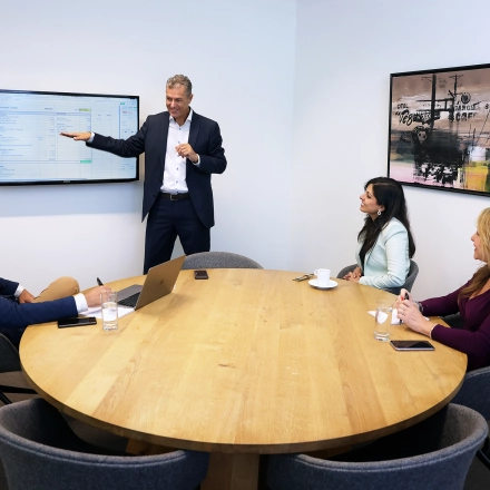 Professionals in a meeting around a wooden table in a furnished office, discussing information displayed on a wall-mounted screen at Koninginnegracht 19, ideal for office space rental.