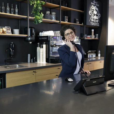A reception area at President Kennedylaan 19 in The Hague Statenkwartier, featuring a smiling receptionist on the phone, modern furniture, and a well-stocked counter. Ideal office space rental.