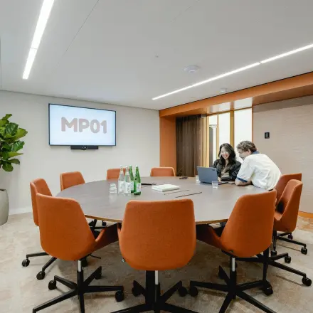 A modern conference room at Moreelsepark 1, Utrecht Central Station with a round table, orange chairs, a large plant, and two people discussing a project. Ideal for office space rental.