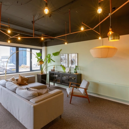 A well-lit office lounge area with modern lighting, a gray sofa, armchair, and decor. Ideal for office space rental at Atoomweg 400, Utrecht Lage Weide.