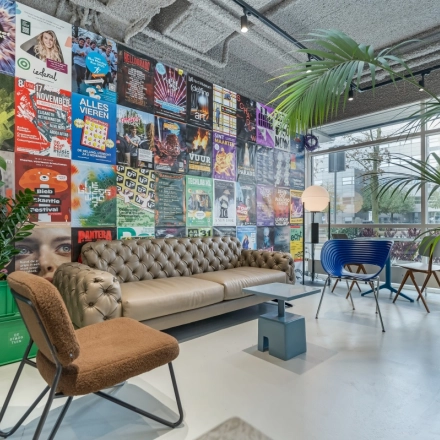 Lively office lounge at Daltonlaan 200, Utrecht Rijnsweerd, featuring colorful posters, a comfortable sofa, stylish chairs, and lush greenery, ideal for informal meetings. A unique twist for office space rental.