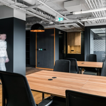 Modern office space with wooden desks and black chairs, featuring exposed ceilings and contemporary lighting at Konstruktorska 11, Warsaw Mokotow. A person walks through the workspace.