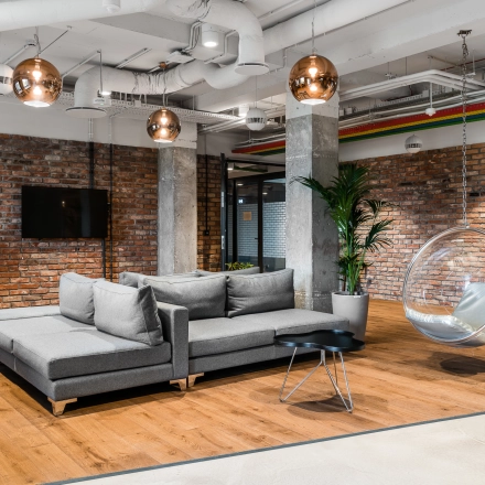 Modern office lounge at Konstruktorska 11, Warsaw Mokotow, with sleek furnishings, brick walls, and a hanging chair, available for office space rental.
