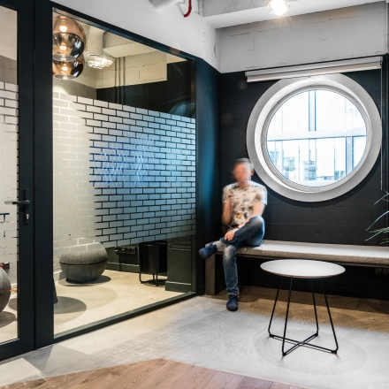 A cozy furnished office space with a round window and seating area at Konstruktorska 11, Warsaw Mokotow. A person is seated, engaging in light business activities.