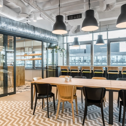 Modern furnished office space at Konstruktorska 11, Warsaw Mokotow with a communal table, high chairs by large windows, and contemporary lighting. Ideal office space rental for collaboration and creativity.