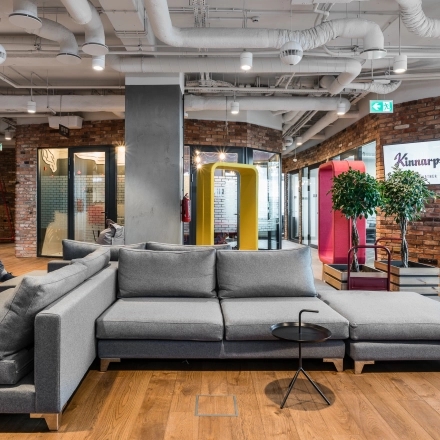 A modern furnished office on Konstruktorska 11, Warsaw Mokotow, featuring a gray sectional sofa and people engaged in conversation, highlighting an inviting space for office space rental.