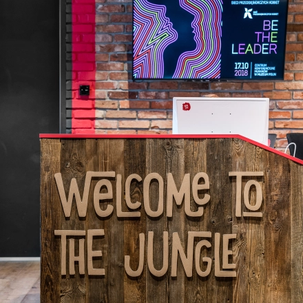 Reception area at Konstruktorska 11, Warsaw Mokotow, ideal for office space rental, featuring a wooden desk with 'Welcome to the Jungle' sign and a wall-mounted screen.