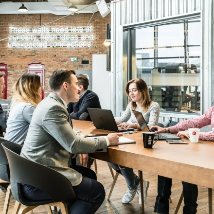Group of people engaged in a meeting around a large wooden table in a modern office space at Al. Jerozolimskie 181B, Warsaw Ochota, available for office space rental.