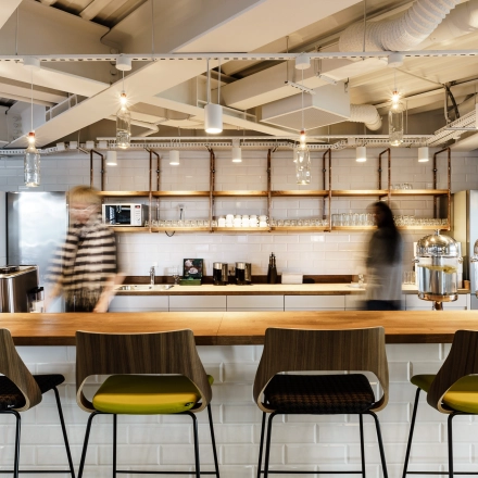 Modern office space in Warsaw Ochota, featuring a furnished kitchen area with sleek lighting and seating. Two individuals are engaging in work-related activities. Ideal for office space rental.