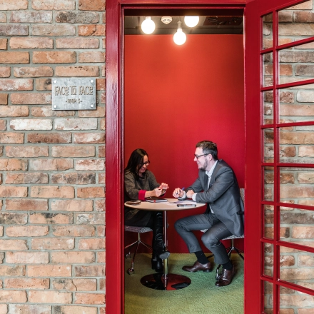 Two people are having a business meeting inside a unique telephone booth-style office space with red walls and a small table.