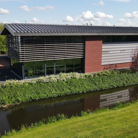 Exterior view of a modern office building for rent office space located at M.A. Reinaldaweg 2d, Woerden. The structure features sleek lines with large glass windows and a landscaped setting beside a canal.