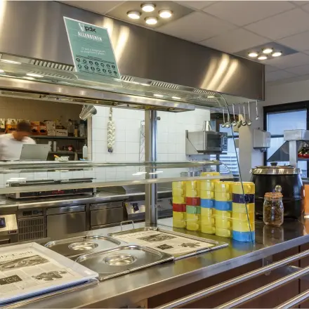 A furnished office pantry with a serving counter, stacked plates, and utensils, located at Bredewater 26, Zoetermeer. An individual behind the counter prepares food.