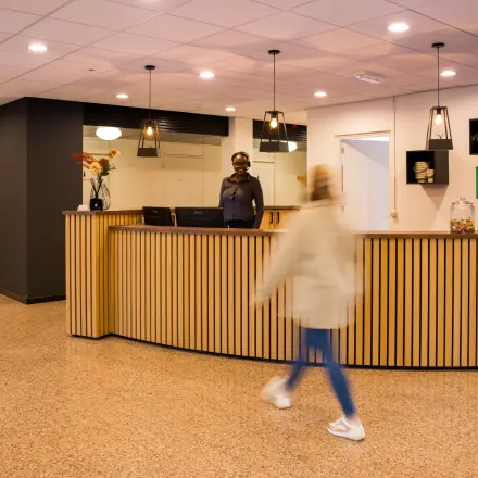Kantoorruimte huren bij Markerkant 1310, Almere Stad. Moderne balie met verlicht plafond en houten details. Medewerker aan receptie, persoon loopt langs. Kantoorpand voor huur.