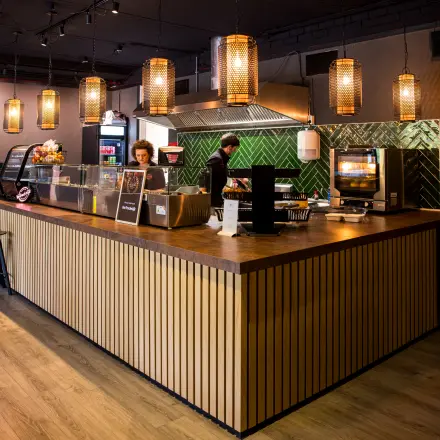 Sfeervolle kantoorruimte huren op Markerkant 1310 in Almere Stad, met een uitnodigende lounge sfeer en houten elementen. Twee mensen zijn bezig met bereiden van drankjes aan de bar.