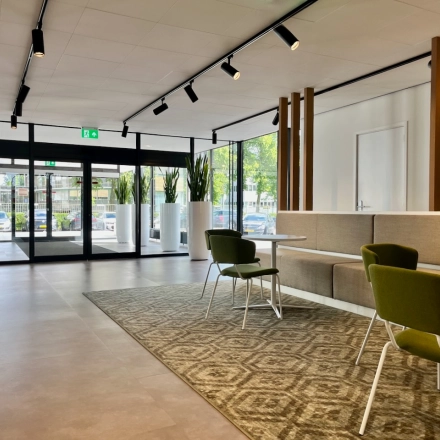 Ingang van een kantoorpand in Amstelveen met moderne inrichting, groene stoelen en grote ramen die natuurlijk licht binnenlaten.