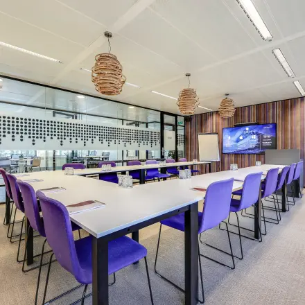 Vergaderruimte met paarse stoelen en grote tafels in een modern kantoorgebouw aan Prins Bernhardplein 200, Amsterdam Amstel. Ruime opzet met scherm en stijlvolle verlichting.