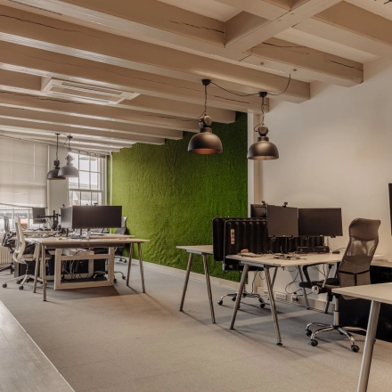 Kantoorruimte huren op Prins Hendrikkade 14 in Amsterdam Centrum met moderne werkplekken en groene muur. De ruimte is gemeubileerd met bureaus en stoelen, ideaal voor zakelijke activiteiten in een kantoorpand.