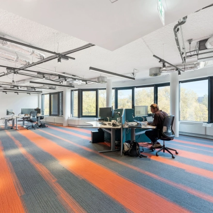 Kantoorruimte te huur op Stadhouderskade 55 in Amsterdam Centrum met een open indeling, grote ramen en kleurrijke vloerbedekking. Een persoon is aan het werk op een volledig ingericht bureau.