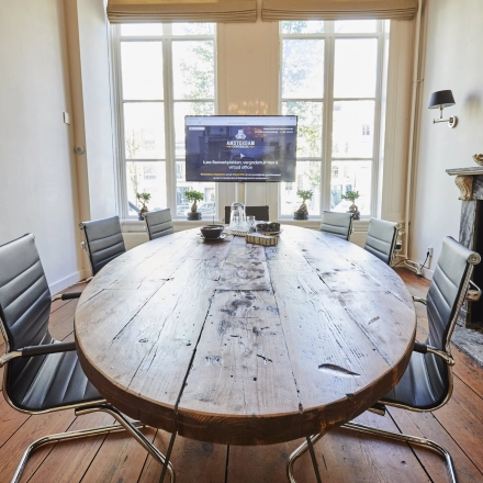 Kantoorruimte huren aan de Keizersgracht 391A in Amsterdam Grachtengordel met een authentieke vergadertafel, stoelen rondom en een flatscreen. De ruimte heeft een karakteristieke houten vloer en grote ramen.