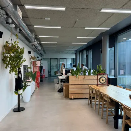 Kantoorruimte huren aan de Moermanskkade 303 in Amsterdam Houthavens, met houten tafels, planten en zicht op lange werkplekken waar mensen werken aan bureaus.