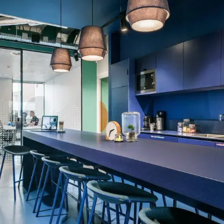 Moderne kantoorruimte aan de Jan van Galenstraat 335 in Amsterdam Nieuw-West, met een grote blauwe tafel en barkrukken, zicht op vergaderruimte. Twee mensen werken achter hun laptops.