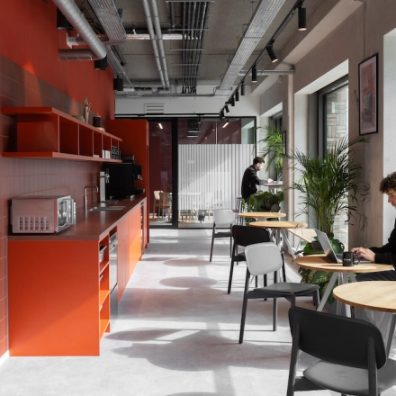 Moderne kantoorruimte huren in Amsterdam Noord met felrode keukenwand, houten tafels, en stoelen. Personen werken op laptops aan tafels bij grote ramen met veel natuurlijk licht.