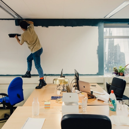 Een kantoor in Amsterdam Noord, TT Vasumweg 44, met een persoon die een muur schildert en bureaus die laptops, kantoorspullen en planten bevatten.