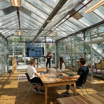 Kantoor met glazen wanden in Amsterdam Riekerpolder, gelegen op Johan Huizingalaan 400. Drie mensen bij vergadertafel, één presenteert aan een scherm. Ruimte biedt een heldere en moderne werkomgeving.