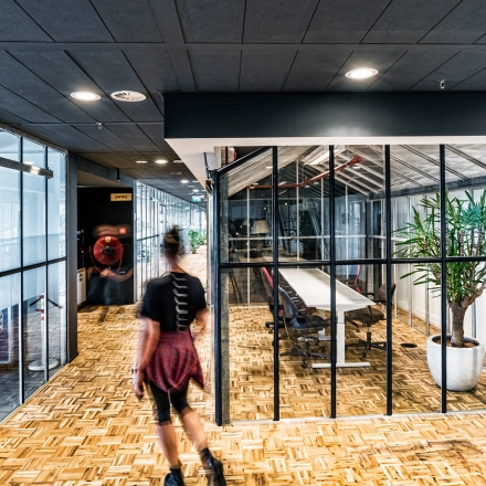 Modern kantoorpand gelegen op Johan Huizingalaan 400, Amsterdam Riekerpolder, met glazen vergaderruimte en houten vloer. Een persoon loopt door de ruimte. Ideaal voor bedrijven die kantoorruimte willen huren.