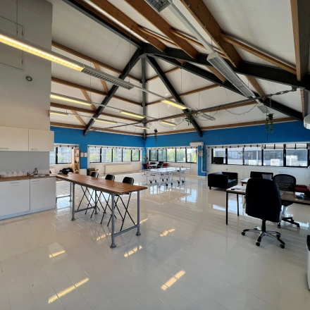 Ruime kantoorruimte huren in Amsterdam Sloterdijk op Dynamostraat 42, met veel natuurlijk licht en houten balken langs het plafond. Verschillende werkplekken en een kitchenette zijn zichtbaar.