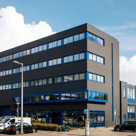 Kantoorpand aan de Kabelweg 24 in Amsterdam Sloterdijk met grijze gevel en blauwe raamkozijnen. Verschillende voertuigen geparkeerd op de parkeerplaats voor het gebouw.