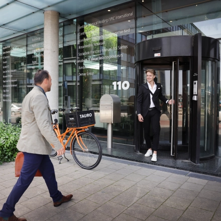 Twee personen voor het kantoorpand op Teleportboulevard 110 in Amsterdam Sloterdijk, een persoon komt naar buiten terwijl een ander met een fiets richting de ingang loopt.