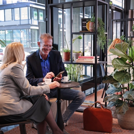 Zakelijke ontmoeting in gemeubileerde kantoorruimte op Teleportboulevard 110, Amsterdam Sloterdijk. Twee mensen overleggen met koffie, omringd door planten en moderne meubels.