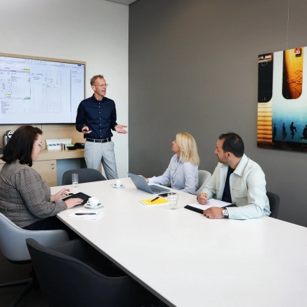 Zakelijke bijeenkomst in een moderne vergaderruimte op Teleportboulevard 110, Amsterdam Sloterdijk. Vier personen, waaronder een presentator, bespreken werkdocumenten bij een lange tafel met laptops en scherm. Kantoor huren mogelijk.