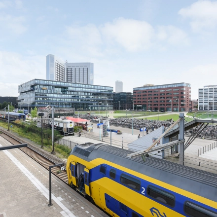 Buitenzijde van kantoorpand op Teleportboulevard 110 in Amsterdam Sloterdijk, met nabijgelegen treinsporen en een gele NS-trein. Ideale locatie voor kantoorruimte huren met uitstekende openbaar vervoer verbindingen.